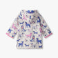 The Hatley Baby Girls’ and Girls’ Little Pups Rain Coat is a children’s hooded rain jacket with a soft terry towelling lining for added comfort. It features playful pink trim and an all-over print of blue, purple, and pink cartoon dogs on a white background.