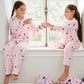 Two children, both dressed in the Hatley Girls’ Night Sky Relaxed Fit Pajama Set featuring matching star and moon designs, sit together on a window seat, smiling and sharing a cloth, with a pile of soft toys and blankets by their side. The pajamas’ soft brushed rib fabric adds to the cozy comfort of the moment.