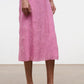A person wearing a pink, knee-length textured skirt, green top, and silver Carrie Sandal by Staud stands on a tan floor against a plain white background.
