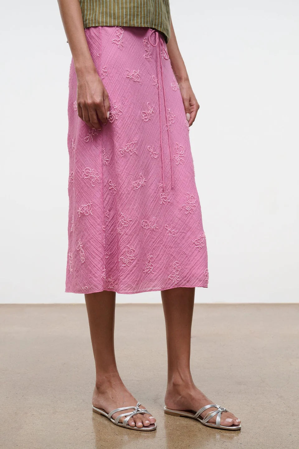 A person wearing a pink, knee-length textured skirt, green top, and silver Carrie Sandal by Staud stands on a tan floor against a plain white background.