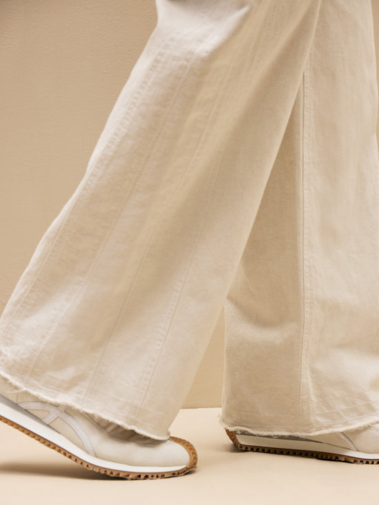 Person wearing TWP Puddle Pant by TWP paired with white sneakers featuring beige-colored soles walks across a beige backdrop.