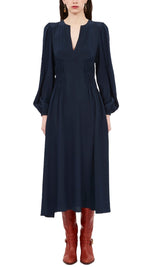 A person wearing the exquisite Ulla Johnson Rouen Dress, crafted from luxurious silk crepe de chine. This long-sleeved, dark blue V-neck design exudes elegance and is beautifully paired with knee-high red boots for a stylish enhancement to its mid-length silhouette.