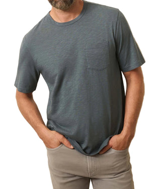 A man wears a short-sleeve, grayish-blue Faherty Sunwashed Pocket Tee made from Regenerative Organic Cotton and light gray pants, standing with his hands in his pockets.