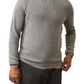 A man wears the Faherty Sunwashed Crewneck Sweater in light gray cotton yarn with dark pants, standing with one hand in his pocket against a white background.
