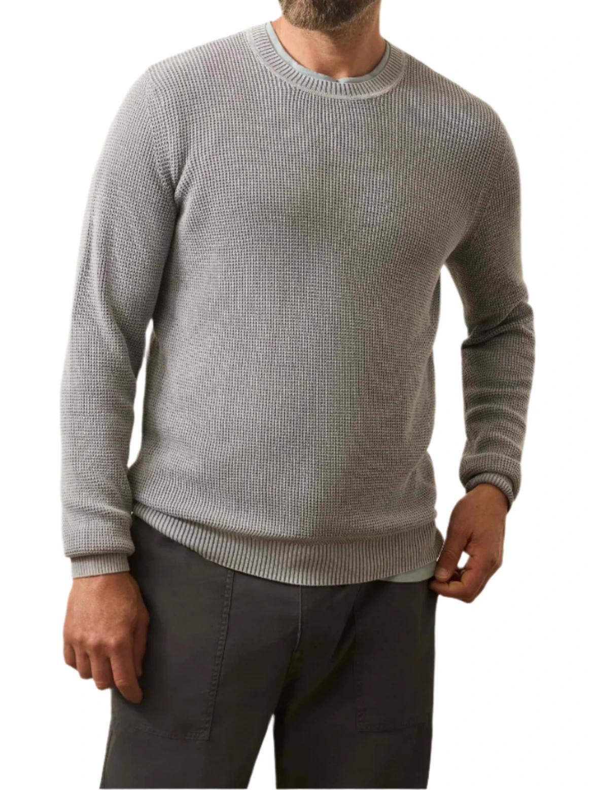 A man wears the Faherty Sunwashed Crewneck Sweater in light gray cotton yarn with dark pants, standing with one hand in his pocket against a white background.