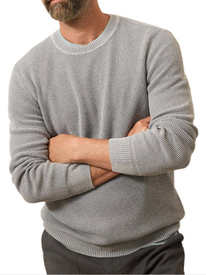 A bearded man stands with arms crossed against a white background, wearing the Faherty Sunwashed Crewneck Sweater in light gray cotton yarn and dark pants.