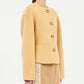 A person wearing the Elsie Wool Coat by Ulla Johnson, a light camel boxy jacket with four large buttons and long sleeves, paired with a beige ruffle-detail skirt.
