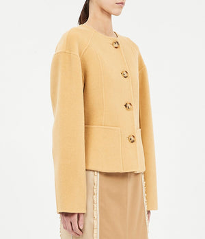 A person wearing the Elsie Wool Coat by Ulla Johnson, a light camel boxy jacket with four large buttons and long sleeves, paired with a beige ruffle-detail skirt.