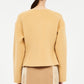 A woman shown from behind wears the Ulla Johnson Elsie Wool Coat, a camel-colored tailored jacket with long sleeves, paired with a beige skirt featuring vertical trim details.