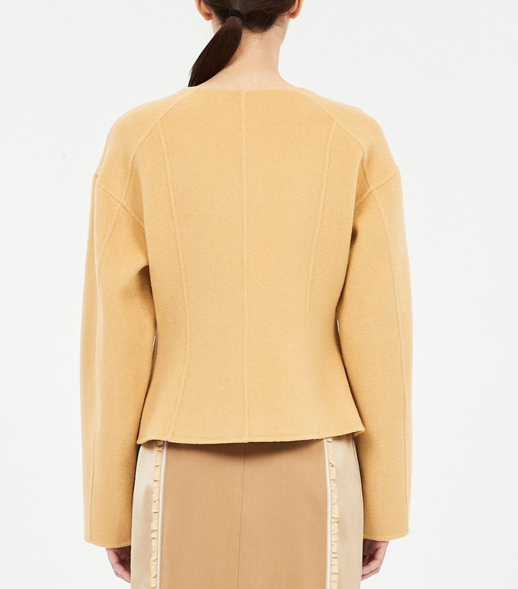 A woman shown from behind wears the Ulla Johnson Elsie Wool Coat, a camel-colored tailored jacket with long sleeves, paired with a beige skirt featuring vertical trim details.