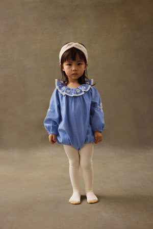 A young child stands facing forward against a plain brown background, wearing a blue long-sleeve romper, white tights, and the Sidony Headband by Louise Misha—a cream-colored knit accessory with fantasy twists and a ruffled floral collar.