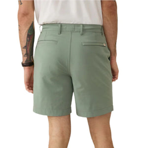 A person is seen from the back in Faherty's light green All Day Short 2.0, 7", paired with a white shirt. A tattoo adorns their right arm as they stand against a plain background, showcasing a casual yet refined style with these tailored shorts.