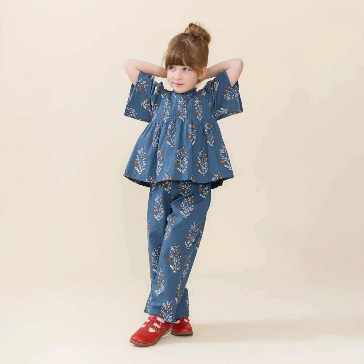 Young girl poses in a blue floral top and Pink Chicken Girls’ Theodore Pant with an elastic waist, paired with red shoes, against a plain beige background.