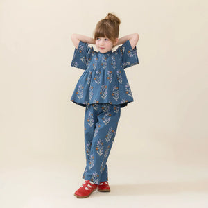 Young girl poses in a blue floral top and Pink Chicken Girls’ Theodore Pant with an elastic waist, paired with red shoes, against a plain beige background.