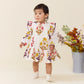A toddler in a Baby Girls’ Harper Dress Set by Pink Chicken with colorful floral patterns stands indoors by a metal vase of assorted dried flowers.