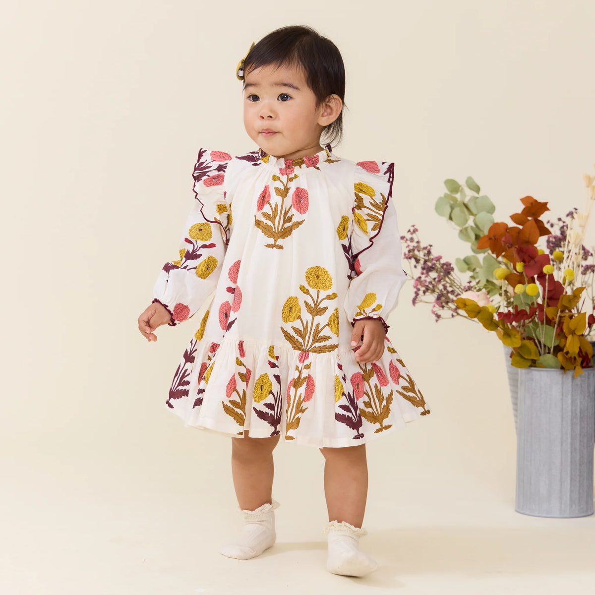 A toddler in a Baby Girls’ Harper Dress Set by Pink Chicken with colorful floral patterns stands indoors by a metal vase of assorted dried flowers.