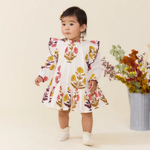 A toddler in a Baby Girls’ Harper Dress Set by Pink Chicken with colorful floral patterns stands indoors by a metal vase of assorted dried flowers.