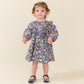 A toddler with light brown hair wears the Pink Chicken Baby Girls’ Everly Dress Set and gray shoes, standing against a plain, light-colored background.
