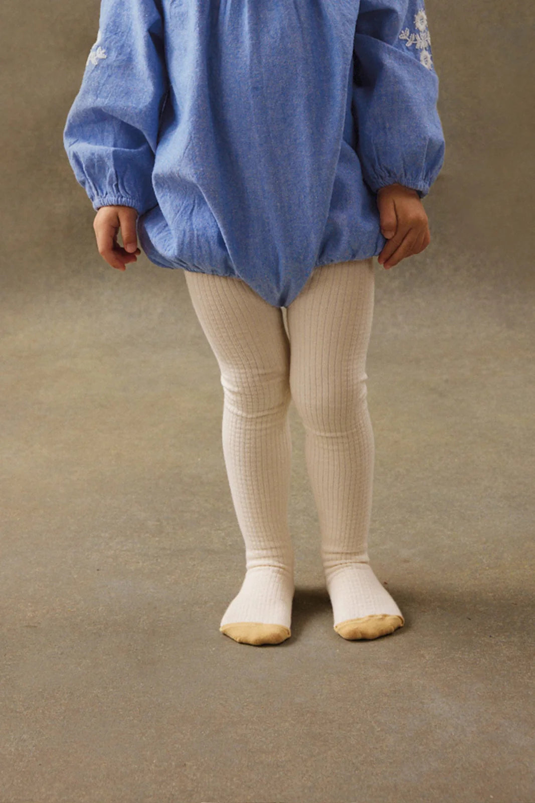 A young child stands on a neutral floor, wearing a blue long-sleeve dress and Louise Misha Baby Girls' and Girls' Chukata Tights in cream-colored soft cotton jersey knit.
