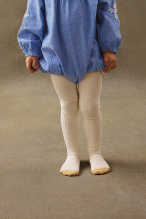 A young child stands on a neutral floor, wearing a blue long-sleeve dress and Louise Misha Baby Girls' and Girls' Chukata Tights in cream-colored soft cotton jersey knit.