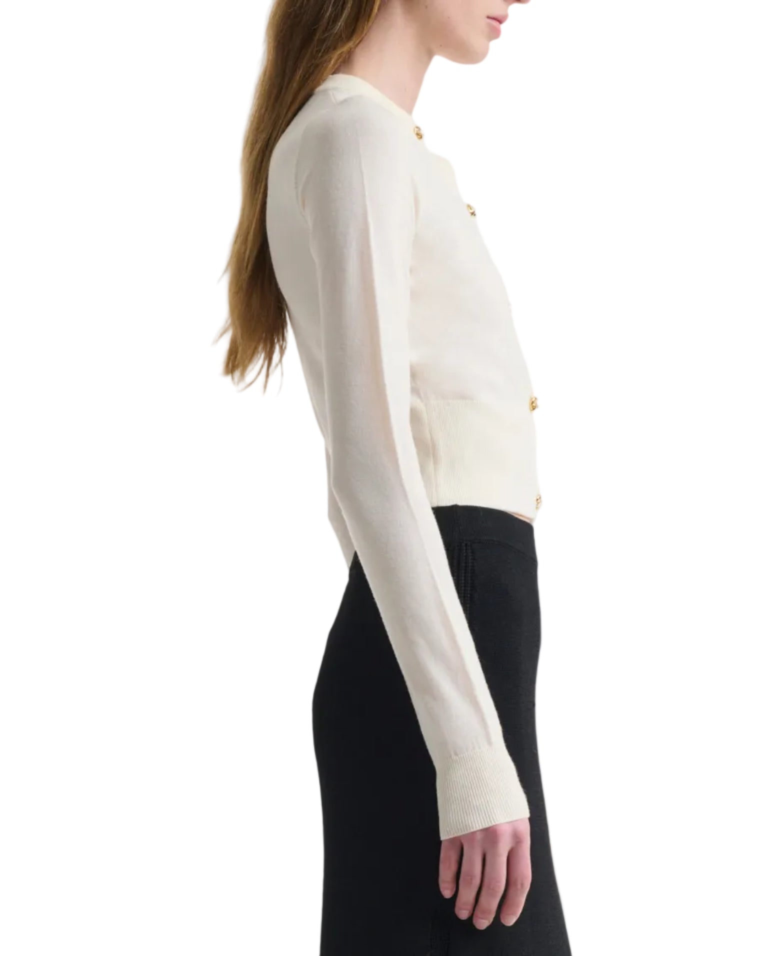A woman wearing the Altuzarra Arlette Sweater—a cream ribbed knit with two-tone metal buttons—paired with a high-waisted black skirt stands sideways against a white backdrop.