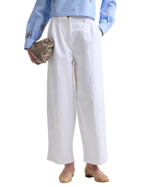 A person wearing white Dash Pant from Altuzarra, a light blue striped shirt, tan flat shoes, and holding a small beige clutch.