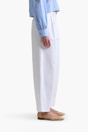 Person standing in profile wearing Altuzarra's white Dash Pant, a blue striped shirt with floral details, and beige flat shoes against a white background—an effortlessly chic look.