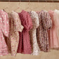 Six Noralee Girls' Louella Dresses in pink and mauve, featuring floral and patterned designs with puff sleeves and tiered skirts, hang on gold hangers from a rod against a beige background.