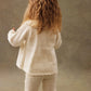 A young child with long wavy hair wears the Louise Misha Baby Girls' Evina Sweater, a cream-colored knit with decorative stitches and matching pants, standing facing away against a neutral background.