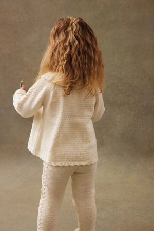 A young child with long wavy hair wears the Louise Misha Baby Girls' Evina Sweater, a cream-colored knit with decorative stitches and matching pants, standing facing away against a neutral background.