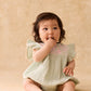 A baby with dark hair sits on the floor wearing the Louise Misha Baby Girls’ Nusha Romper in light green cotton gauze with pink embroidery, looking up with fingers in their mouth against a neutral beige background.