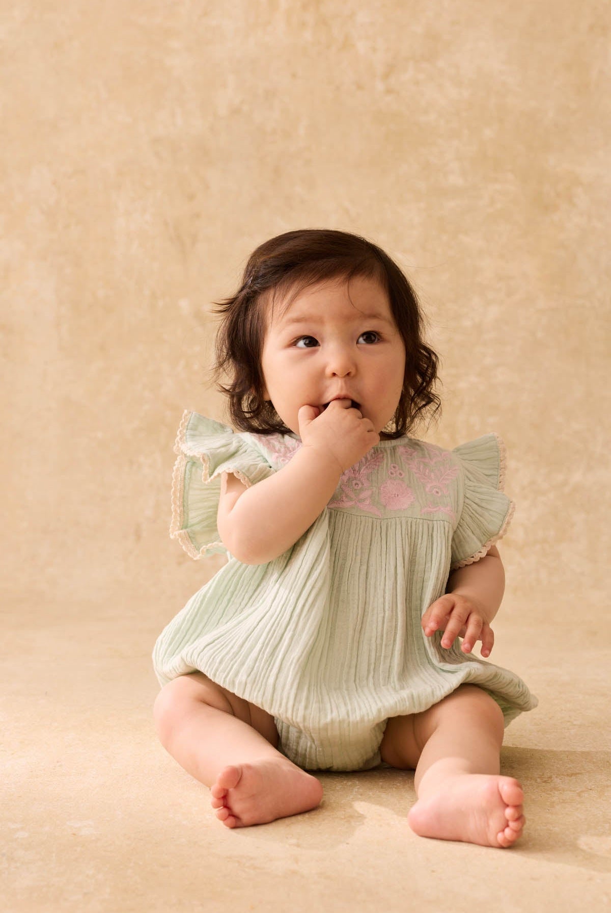 A baby with dark hair sits on the floor wearing the Louise Misha Baby Girls’ Nusha Romper in light green cotton gauze with pink embroidery, looking up with fingers in their mouth against a neutral beige background.