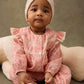 A baby girl in the Louise Misha Lilany Jumpsuit, featuring pink cotton batiste with ruffled armholes and lace details, and a cream headband, sits before a large plush toy and looks toward the camera.