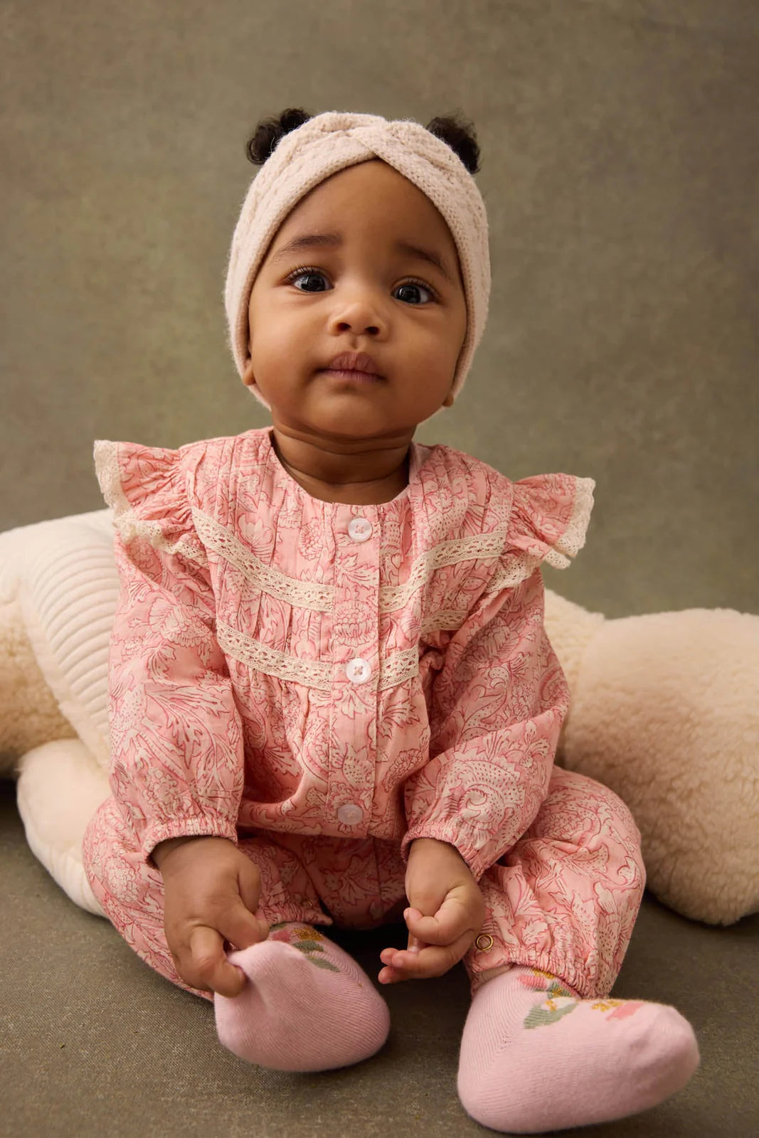 A baby girl in the Louise Misha Lilany Jumpsuit, featuring pink cotton batiste with ruffled armholes and lace details, and a cream headband, sits before a large plush toy and looks toward the camera.