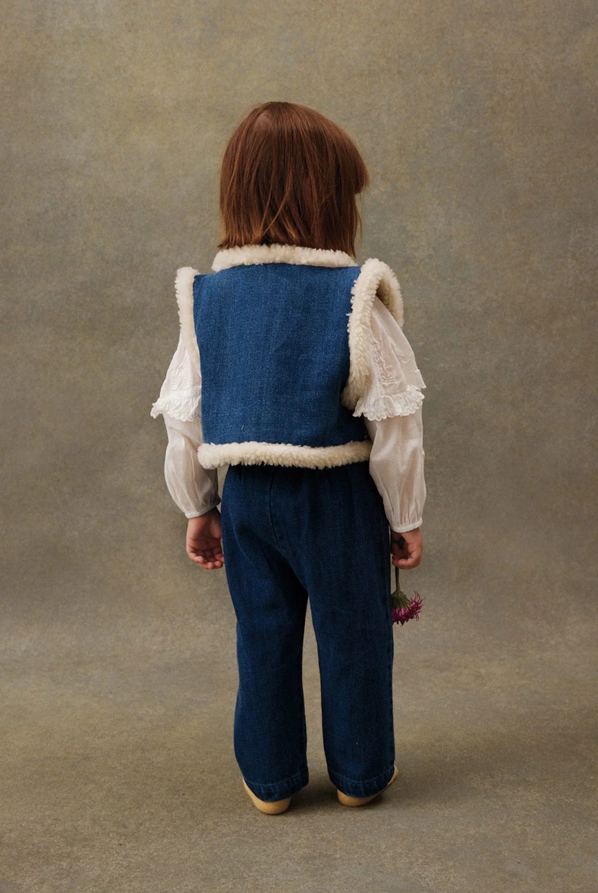 A young child faces away, wearing Louise Misha's Baby Girls' Flofia Pants with an elastic waistband, a blue denim vest trimmed in white, a white long-sleeve blouse, and holding a pink flower in her left hand.