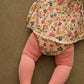A baby wears the Louise Misha Baby Girls' India Romper, featuring a floral print and ruffle collar, lying face down on a brown surface with only her lower body in pink striped tights visible.