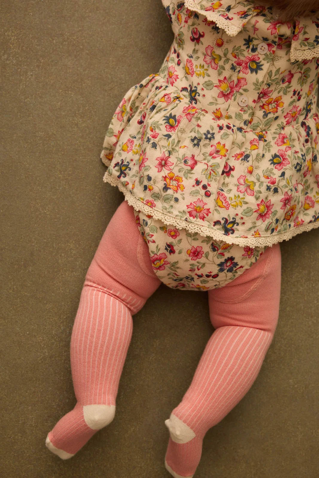 A baby wears the Louise Misha Baby Girls' India Romper, featuring a floral print and ruffle collar, lying face down on a brown surface with only her lower body in pink striped tights visible.