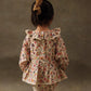 A young child wearing the Louise Misha Baby Girls' India Romper with a floral hair tie and cream tights stands facing away against a neutral, textured backdrop. The romper features a floral print, ruffle collar, and lace edges.