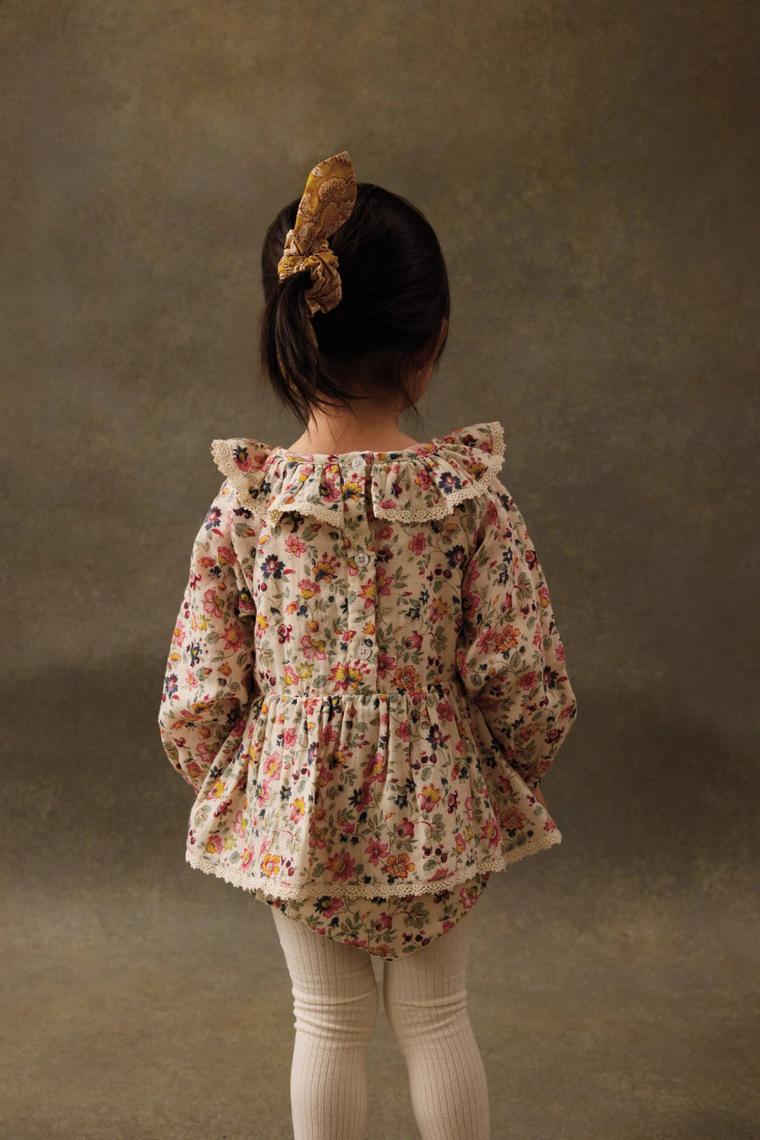 A young child wearing the Louise Misha Baby Girls' India Romper with a floral hair tie and cream tights stands facing away against a neutral, textured backdrop. The romper features a floral print, ruffle collar, and lace edges.