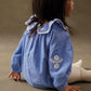 A young child with straight dark hair sits sideways on the floor, wearing the Louise Misha Baby Girls' Ilani Romper in blue chambray with white embroidery and a ruffled collar, paired with cream tights.