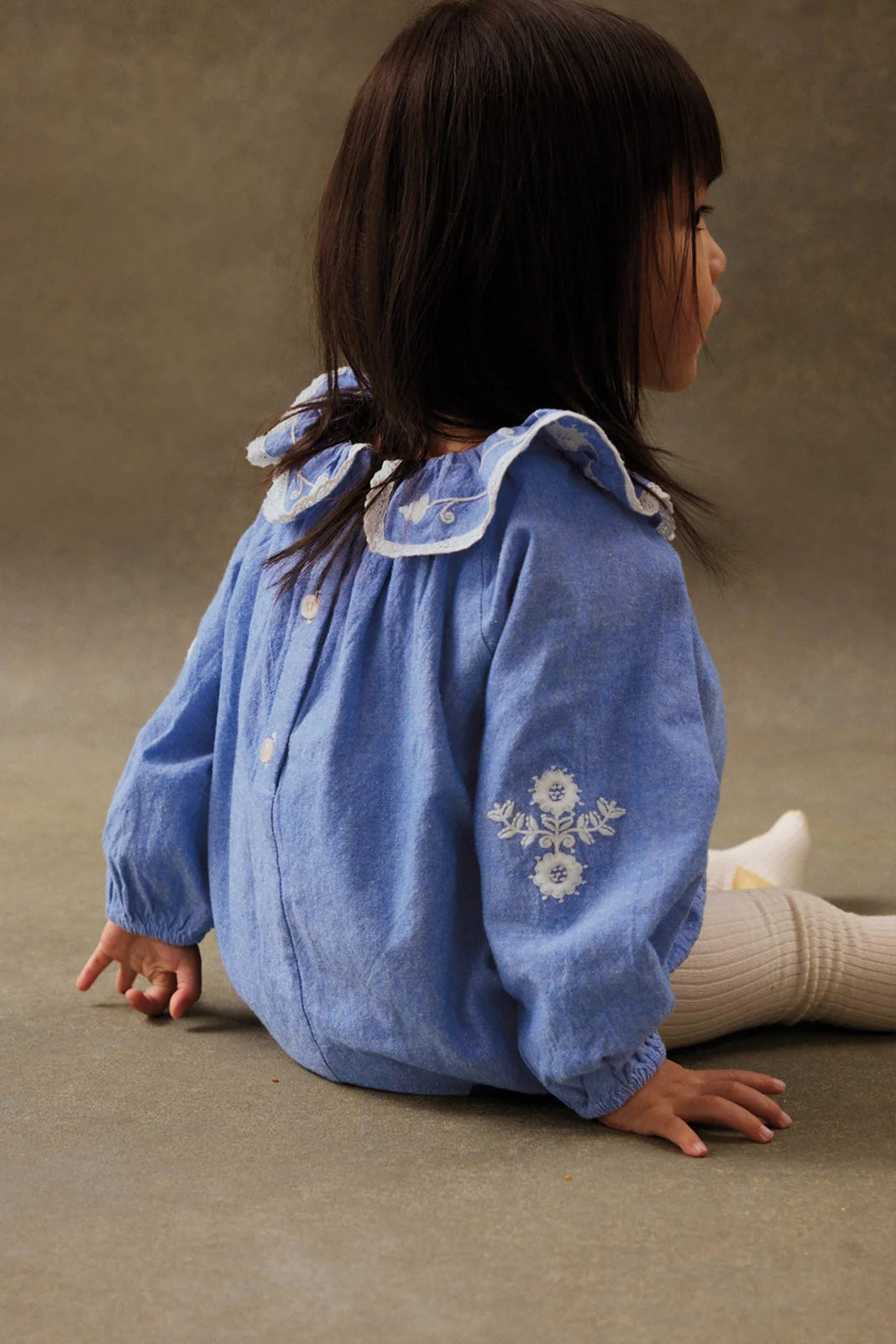 A young child with straight dark hair sits sideways on the floor, wearing the Louise Misha Baby Girls' Ilani Romper in blue chambray with white embroidery and a ruffled collar, paired with cream tights.