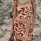 A woman stands against a rocky background, wearing the Johanna Ortiz Raiz Ritual Mini Dress—a brown and white strapless linen mini dress with a wrap-effect skirt—paired with a large shell necklace and a woven straw bag.