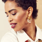A woman with short curly hair wears a white shirt, Lizzie Fortunato Organic Gold Hoops, and a chunky necklace with gold and orange links. She looks down and smiles slightly.