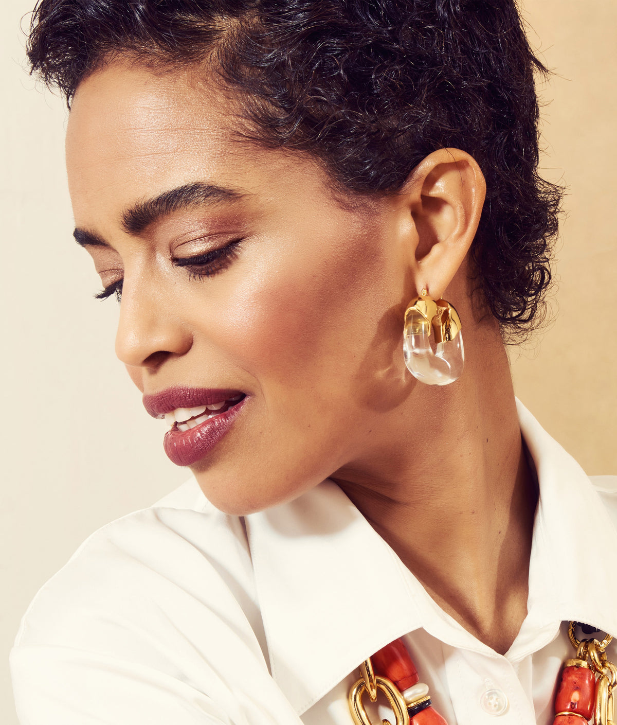 A woman with short curly hair wears a white shirt, Lizzie Fortunato Organic Gold Hoops, and a chunky necklace with gold and orange links. She looks down and smiles slightly.
