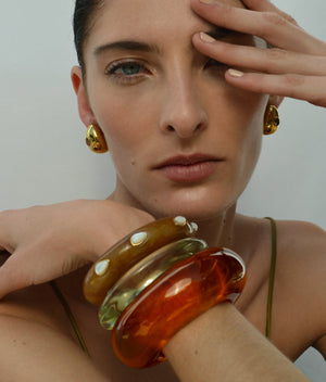 A woman with light skin wears gold earrings and chunky resin bracelets—amber, green, clear with white dots—plus the Lizzie Fortunato Ridge Cuff With Stones, a dark mocha acrylic domed cuff, posing with her hand partly covering her face.