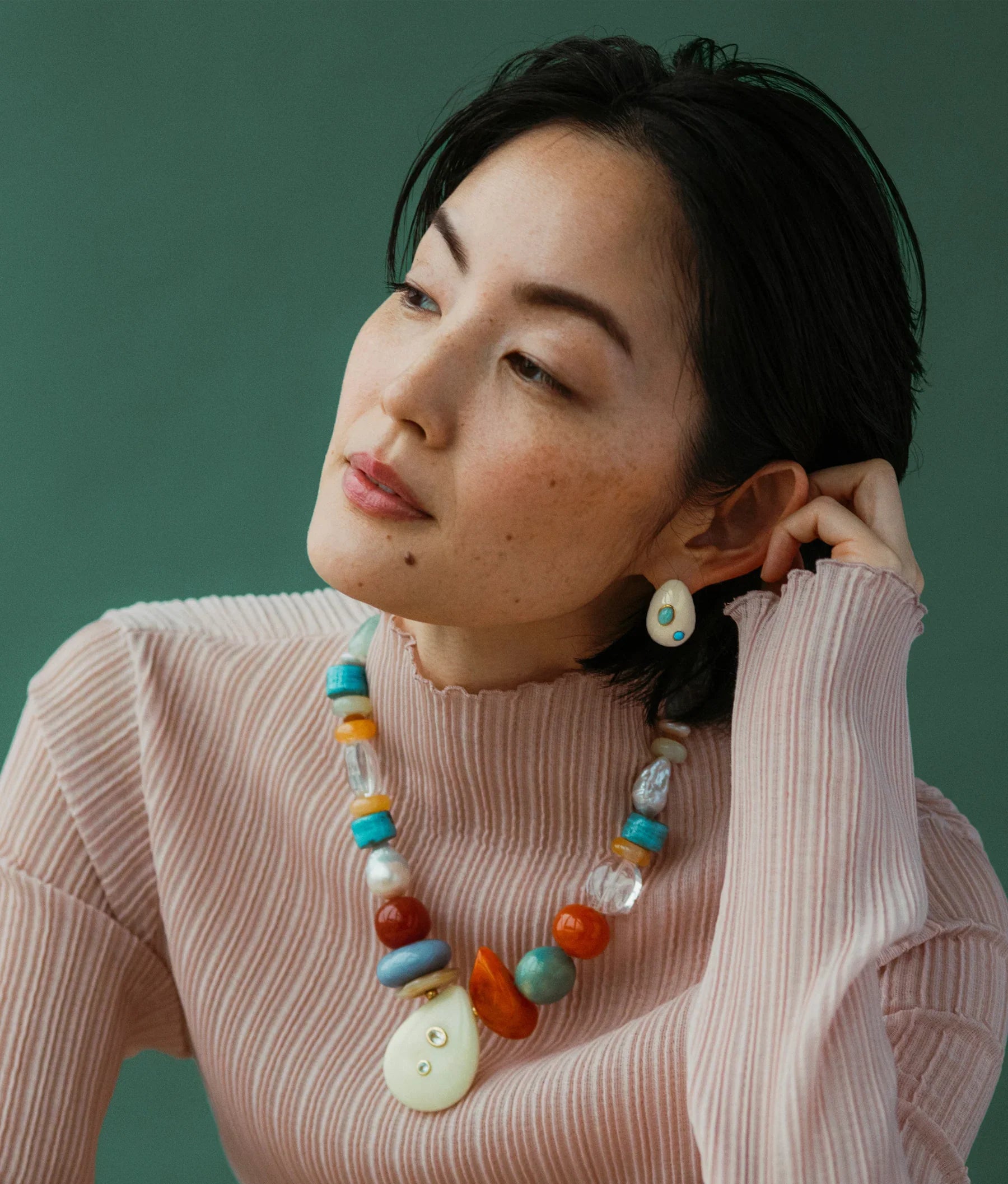 Woman wearing a ribbed, light pink turtleneck and colorful beaded necklace, looking to her left with her hand touching her Lizzie Fortunato Mini Arp Earrings by Lizzie Fortunato. Background is a solid green.