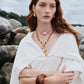 A woman with long red hair wears a white dress, Lizzie Fortunato Organic Mini Gold Hoops, layered necklaces, and a large brown bracelet while standing outdoors in front of large rocks.