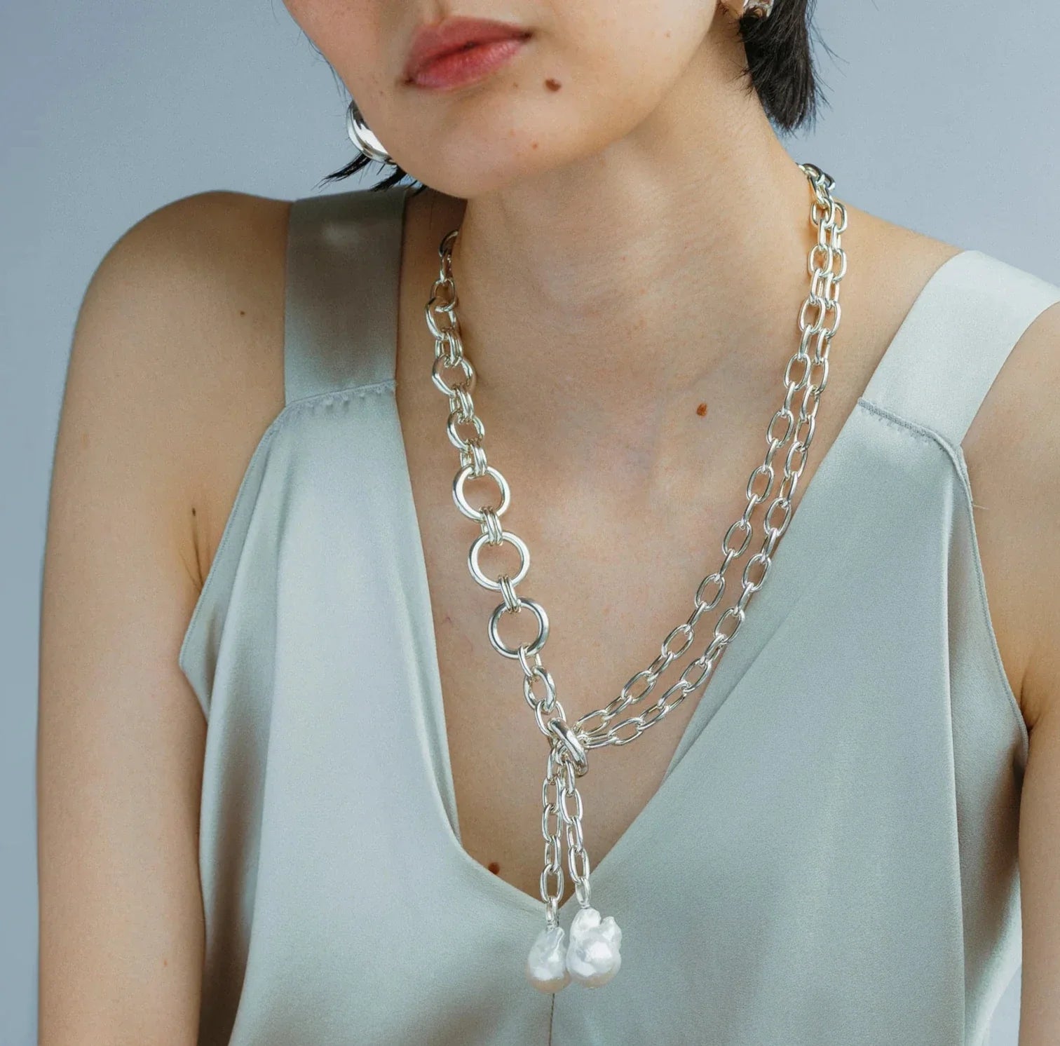 A person elegantly wears the Lizzie Fortunato Edith Lariat, paired with a silky light gray top.