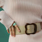 Person wearing a ribbed pink top, the Lizzie Fortunato Florence Belt featuring a large clover-inspired buckle, and a green ring on their finger, posed against a green background. Sleekly chic in every detail.