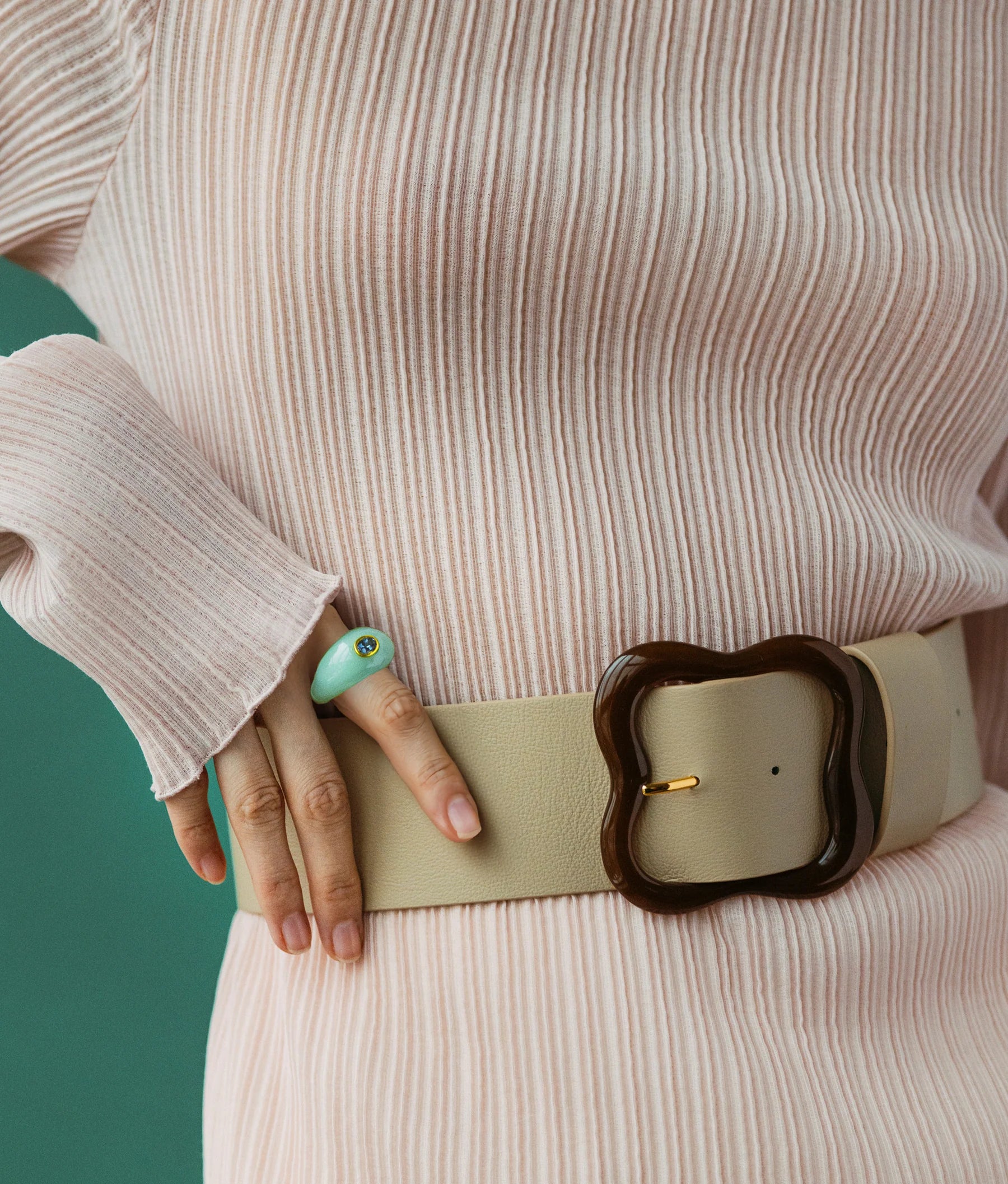 Person wearing a ribbed pink top, the Lizzie Fortunato Florence Belt featuring a large clover-inspired buckle, and a green ring on their finger, posed against a green background. Sleekly chic in every detail.